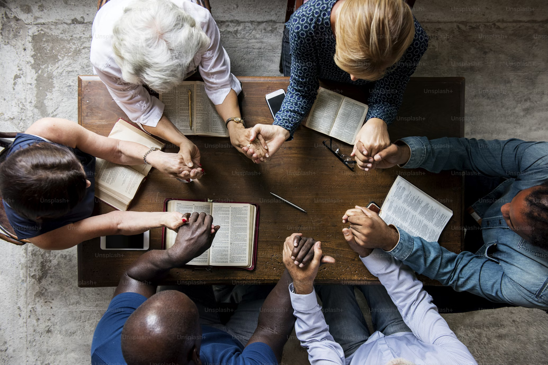 Grupo de seis pessoas sentadas ao redor de uma mesa, com Bíblias abertas à sua frente, unidas de mãos dadas em momento de oração e agradecimento. Cena representa união, fé e estudo bíblico em comunidade.